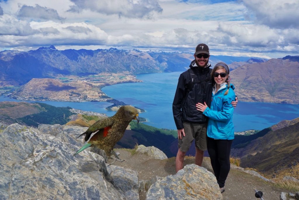 A KUHL Ambassador and a KUHL man standing on a peak with a rocky mountains in the distance, a lake below, and a parrot photo-bombing