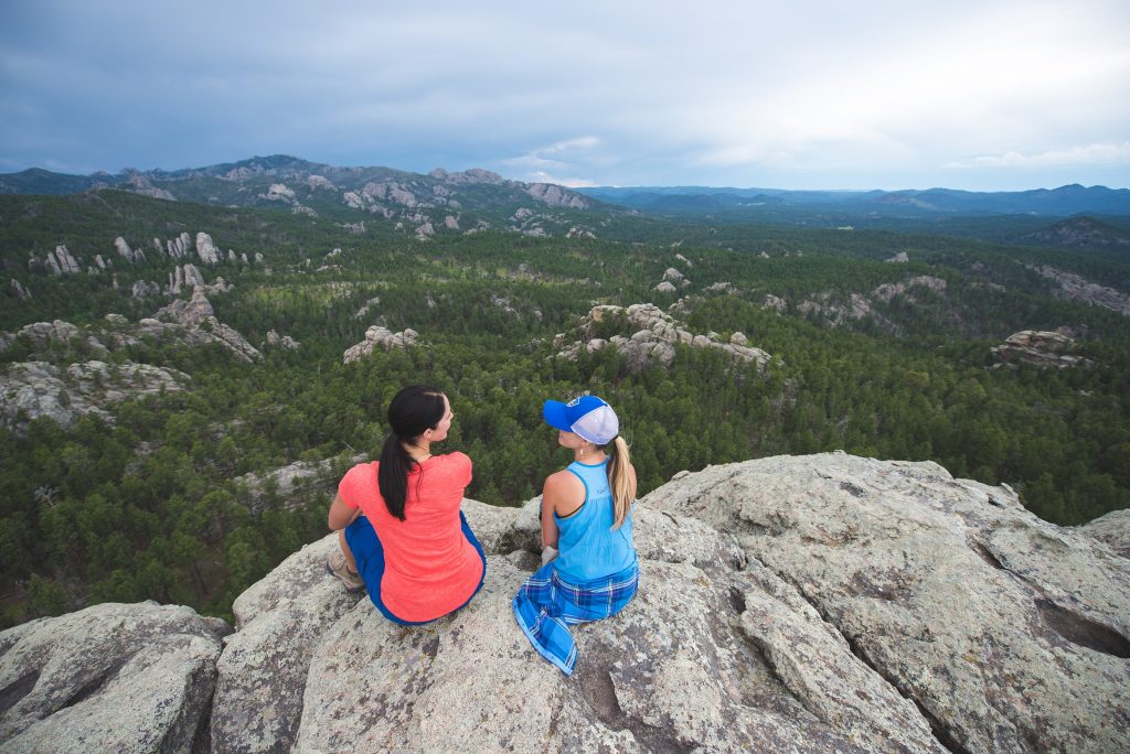 Two women sitting on a rock overlooking a forest, dressed in KUHL clothing.