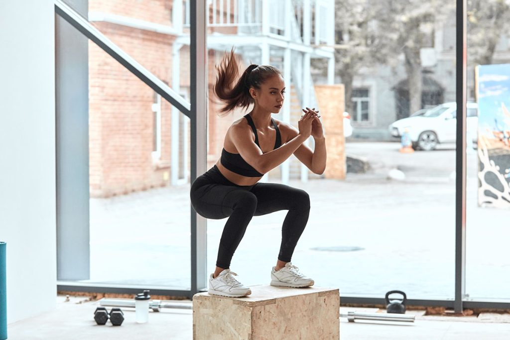Young athletic woman do squats on wooden box in gym
