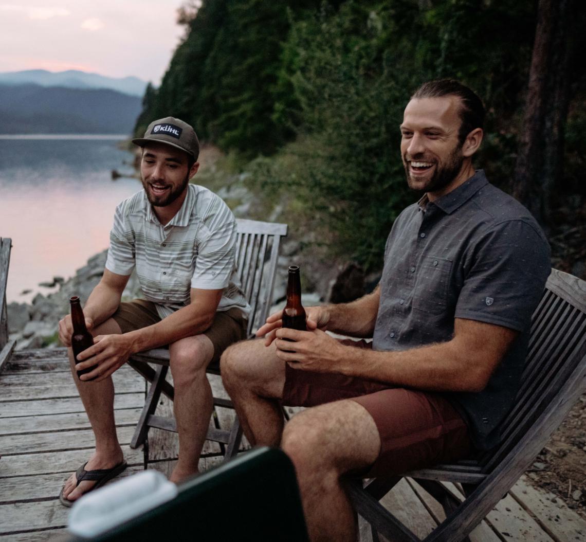 Two men enjoying drinks at a lake wearing Kühl casual shorts