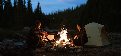 A man and a woman sitting next to a campfire while camping in a pine forest.