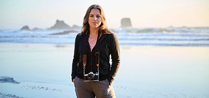 A woman with a camera stands on a sea shore, with rocky cliffs in the background