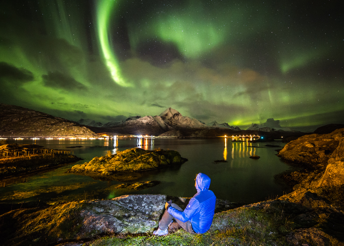 A man in KUHL winter hiking clothing sitting on a rock and looking at northern lights