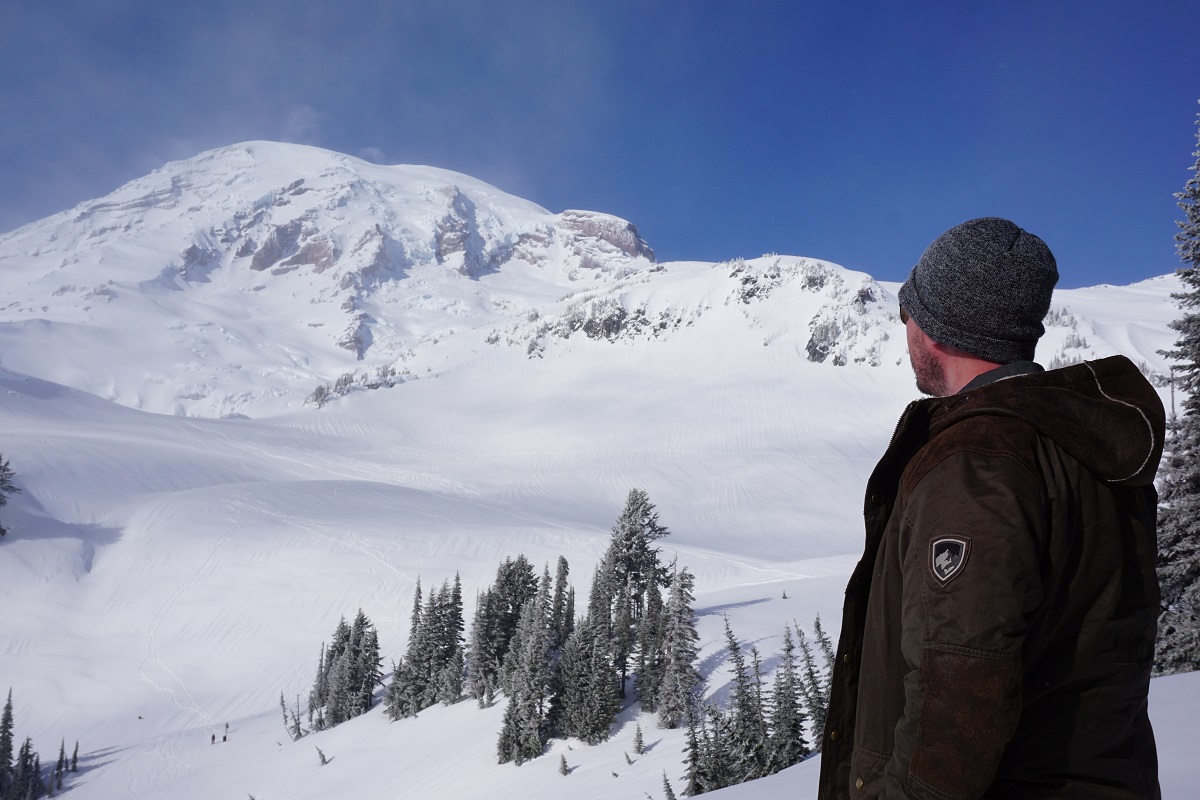 A man standing at a foothill of a snow covered mountain, dressed in KUHL men's winter hiking jacket