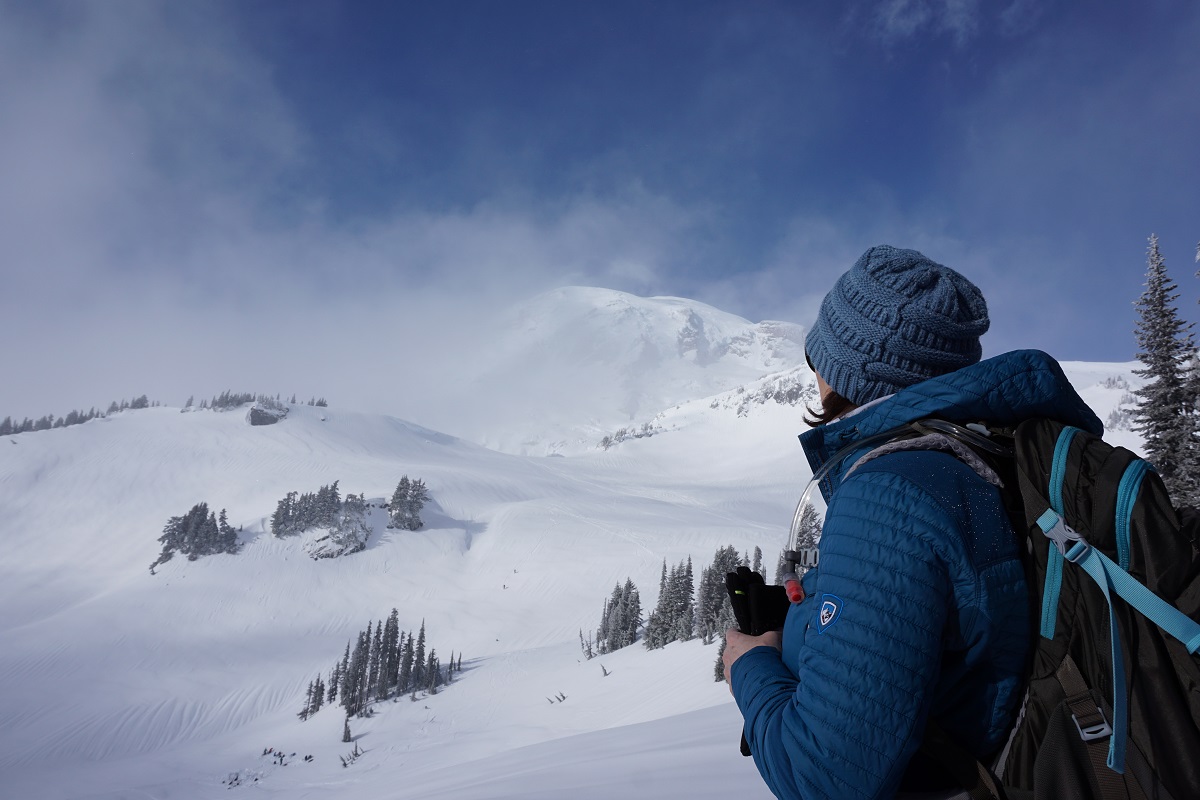 A woman standing at a foothill of a snow covered mountain, dressed in KUHL men's winter hiking jacket