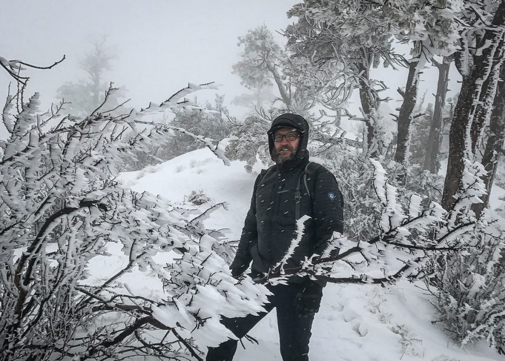 A man standing on a wintery snowy trail dressed in KUHL men's winter hiking jacket