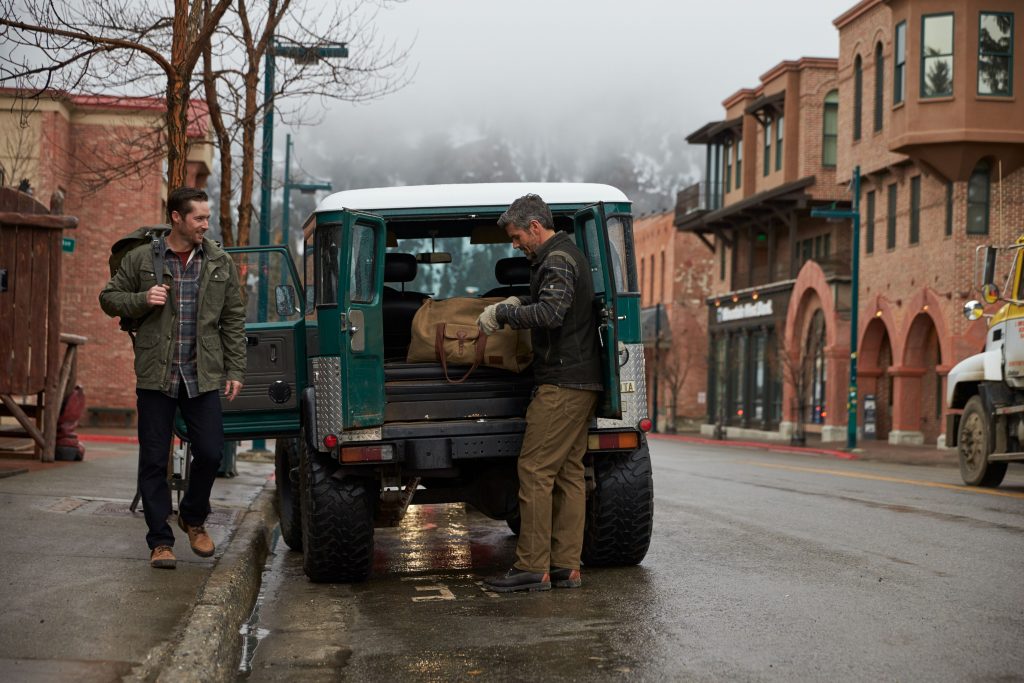 Two men standing next to a teal truck in a snowy small town.