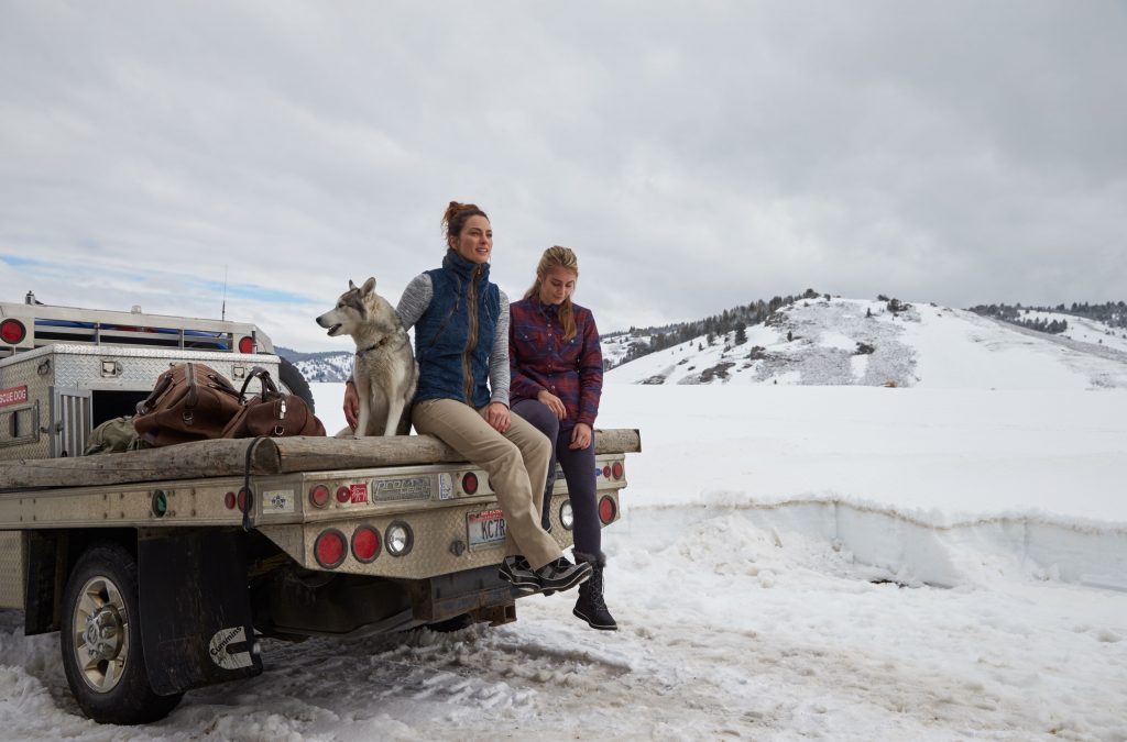 Two women sitting on the back of a truck, petting a dog in a winter scenery.