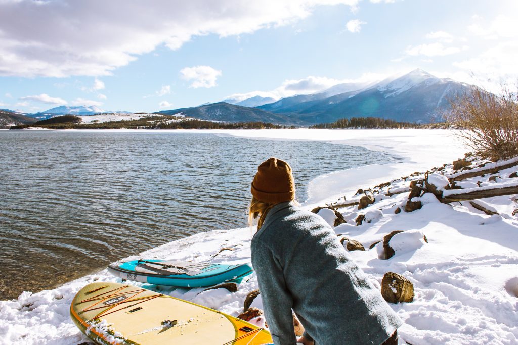A woman pushing her paddling board in a river from a snowy shore.