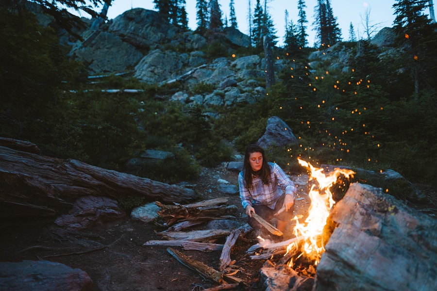 How to Survive Until Help Arrives - A woman in KUHL clothing crouching next to a campfire in the wilderness.