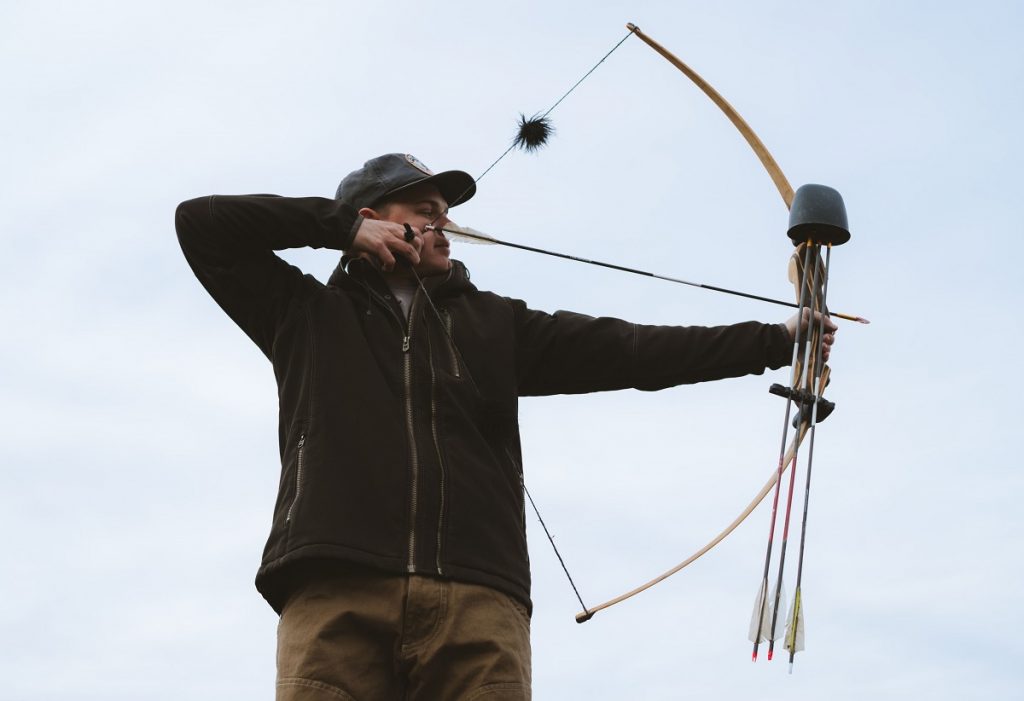 man in KÜHL jacket and KÜHL pants aiming with bow and arrow