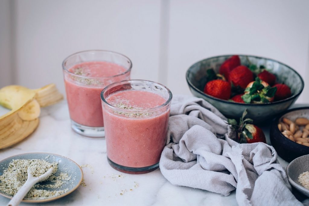 two glasses with pink smoothie next to a bowl of strawberries and banana