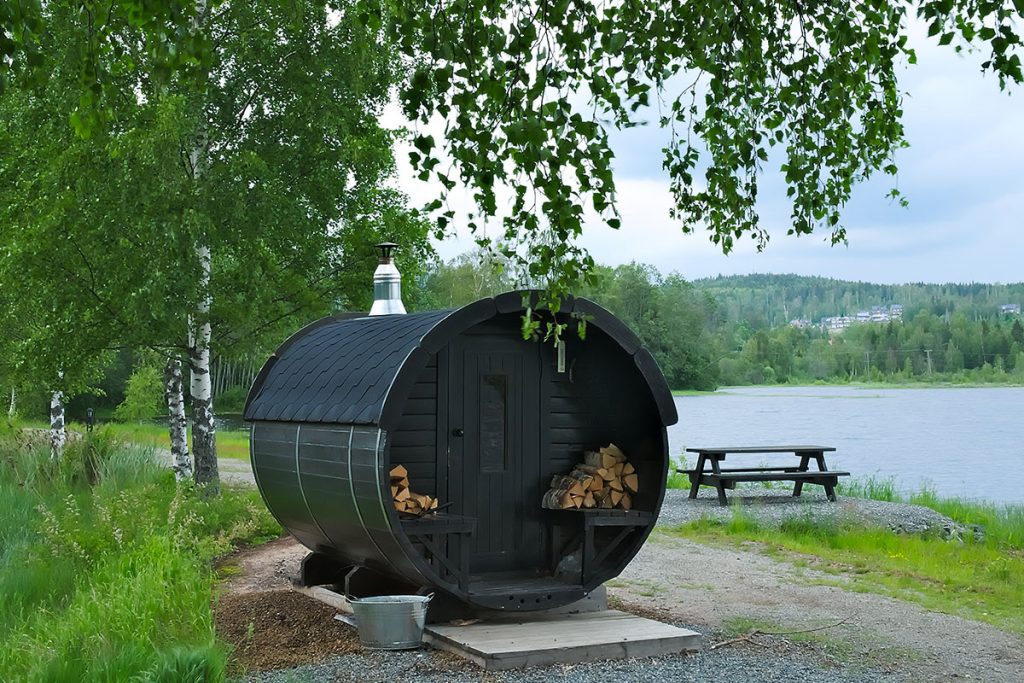 Round wooden outdoor sauna with stacks of wood for heating, by a lake with nearby trees and a picnic table