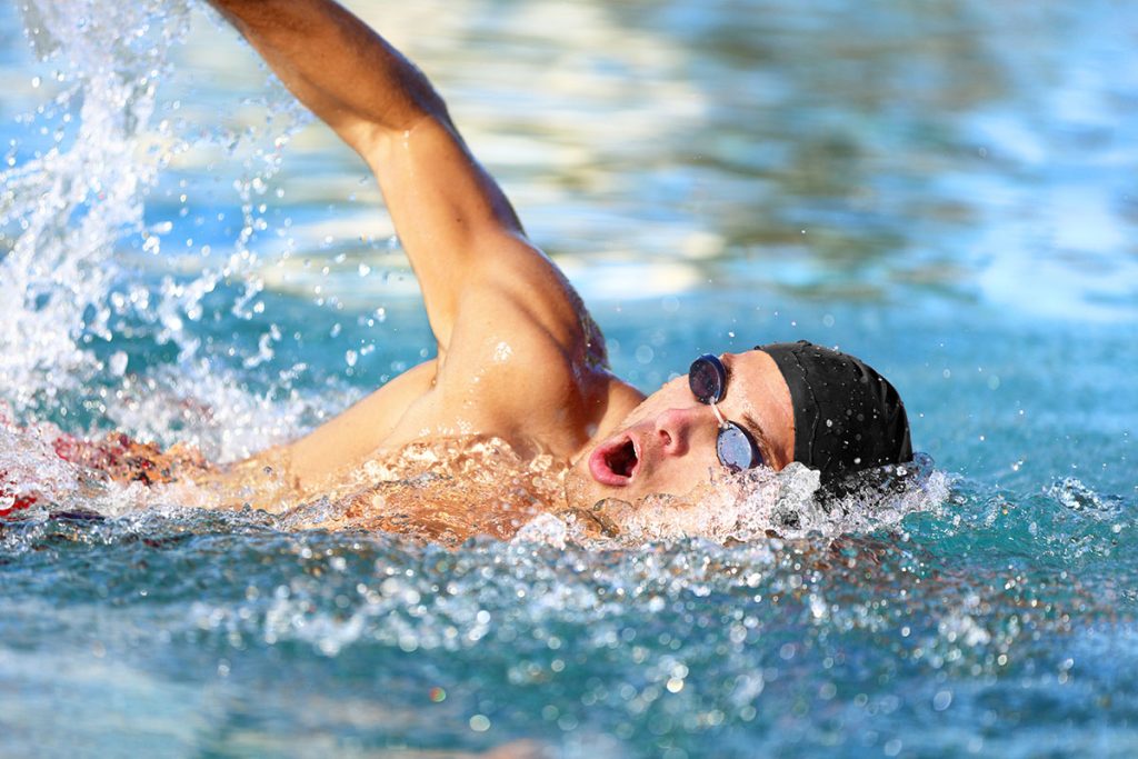 Portrait of an athletic young male triathlete swimming crawl wearing cap and swimming goggles.
