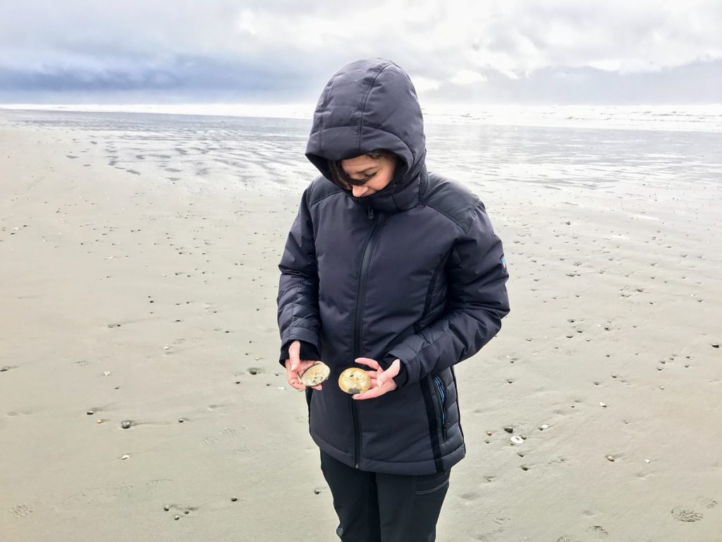 A woman in KUHL women's winter hiking jacket standing on a shore on a cold day, holding sea shells