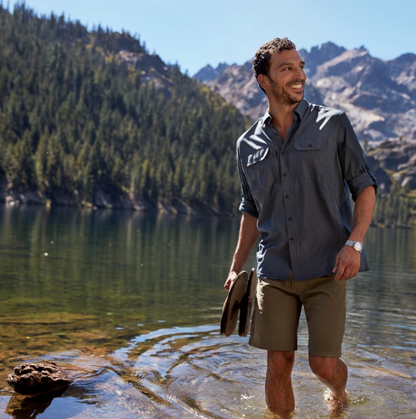 A man walking in a lake holding his flip-flops, mountain and forest in the background