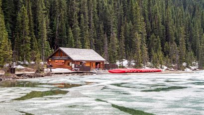 Brown wooden house surrounded by pine trees, kayaks and iced lake
