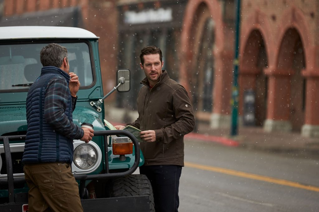 Two men standing next to a teal truck in a snowy small town.