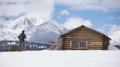 The Magic of Snow: A man standing on a snow next to a wooden cabin, overlooking a mountain