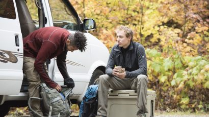 Two men preparing for a hike in a van, one is adjusting his backpack and the other is sitting on a box.