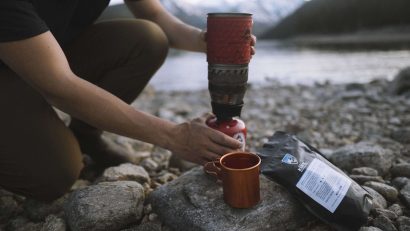 man holding a portable stove kneeling next to brown mug and black bag on rocky ground