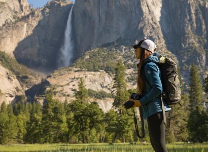 woman in blue KÜHL jacket holding camera in front of mountain with waterfall
