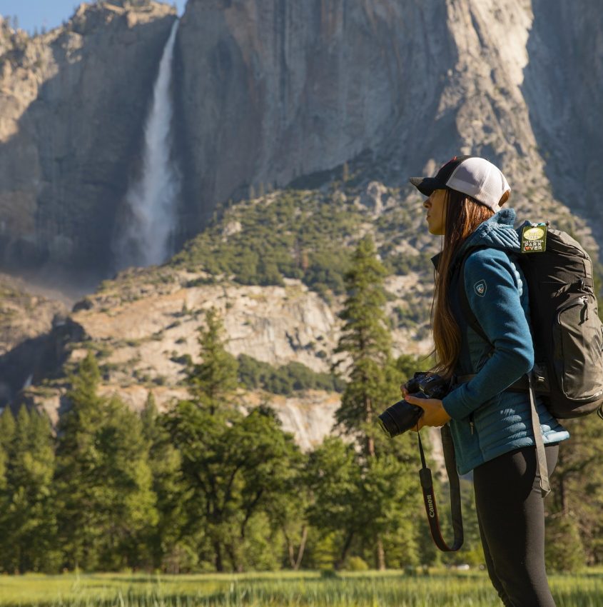 woman in blue KÜHL jacket holding camera in front of mountain with waterfall