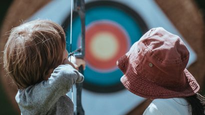 boy aiming bow on target while girl with red hat watches