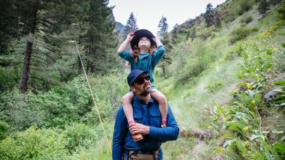 man in KÜHL shirt and KÜHL hat carrying daughter on his shoulders