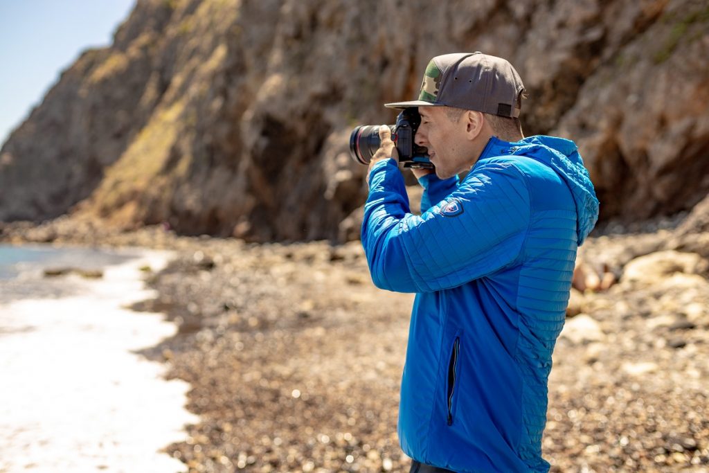 man in blue KÜHL jacket holding camera on coastline