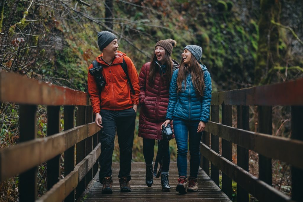 man and two women in KÜHL jacket smiling while walking on brown bridge