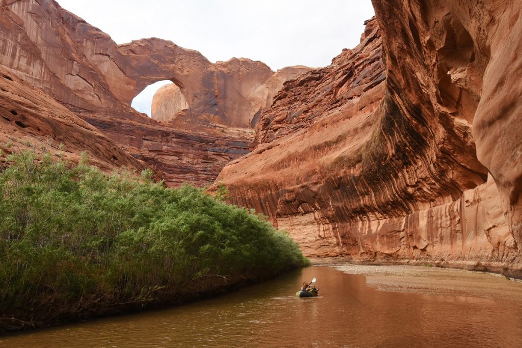 man packrafting on brown river next to rock formations