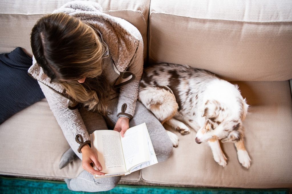 woman in KÜHL Flight Jacket reading book on the couch next to white dog
