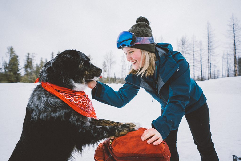 woman in KUHL shell playing with black dog on snowy field
