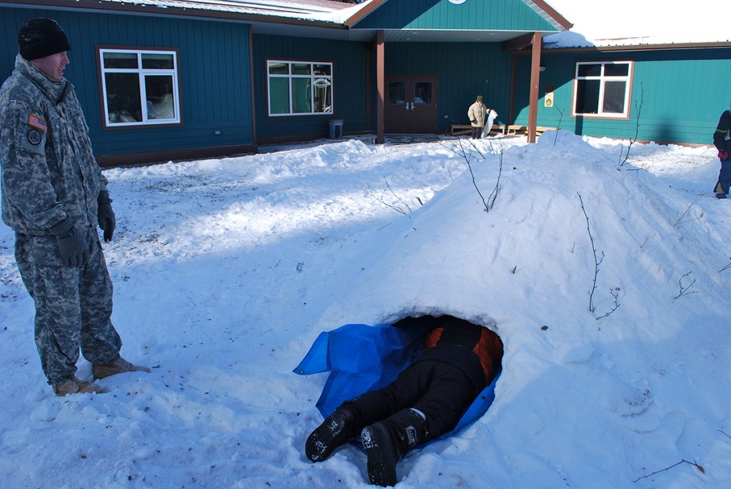 man watching another man laying on blue cloth inside winter survival shelter