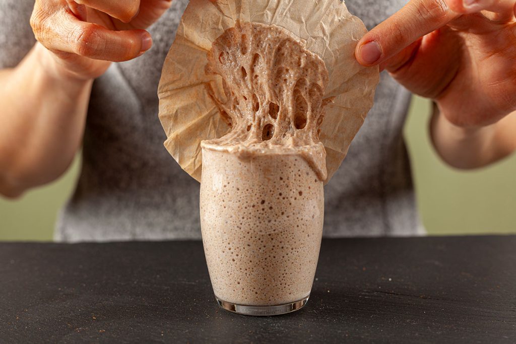 A woman is carefully removing the top cover of a sourdough starter culture which is about to overflow the glass cup