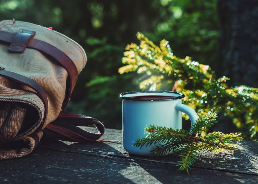 Enameled cup of tea, backpack of traveller on wooden board in summer forest outdoors