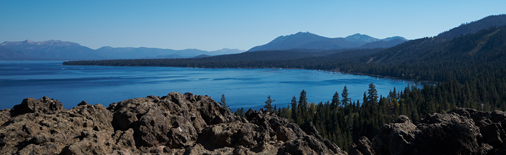 A landscape image of a rocky mountain and pine forest in the distance, surrounding a lake. 